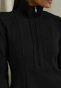 Schwarzer Pullover mit hohem geripptem Kragen und einem halben Reißverschluss, aus glattem Stoff mit Nahtdetails für eine strukturierte Passform.