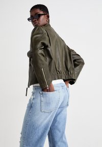 Chaqueta bomber de cuero color verde oliva con dobladillo elástico y cremalleras en las mangas, combinada con jeans desgastados de color azul claro.