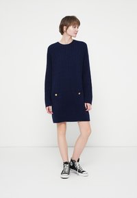 Rochie tricotată bleumarin cu textură canelată, mâneci lungi, două buzunare frontale și accente cu butoane aurii la buzunare.