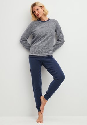 Femme appuyée contre un mur blanc, vêtue d'un haut gris à manches longues et d'un pantalon de jogging bleu marine, pieds nus, un pied levé.