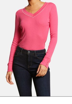 Frau trägt ein hellpinkes langärmeliges V-Ausschnitt-Top mit Knopfdetails an den Ärmeln, kombiniert mit dunkelblauen Jeans.
