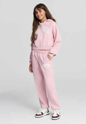 Hoodie cropped rosa con lettering bianco, pantaloni della tuta rosa abbinati con tasche e sneakers bianche con accenti scuri. Tessuto morbido, vestibilità rilassata.