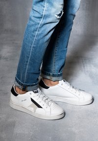 Białe skórzane sneakersy z czarnymi i szarymi akcentami, okrągły nos, perforowane detale i teksturowana gumowa podeszwa, w połączeniu z niebieskimi dżinsami.