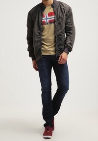 Grå bomberjakke, beige t-shirt med rødt og blåt grafisk design, mørke jeans og røde sneakers. Afslappet outfit med en løstsiddende pasform.