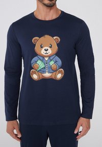 Camiseta de manga larga en color marino con un oso de dibujo animado que lleva una chaqueta azul, sosteniendo billetes, con una cara redonda y detalles en sus patas. Textura de tela suave.