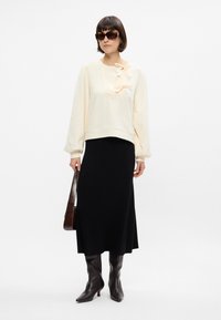 Sweatshirt crème avec des nœuds, jupe midi noire, bottes en cuir marron jusqu'aux genoux et lunettes de soleil en écaille de tortue, se tenant devant un fond blanc.