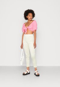 Top cropped rosa com mangas bufantes e laço frontal, combinado com calças justas amarelas claras com padrão xadrez e sandálias brancas com solas pretas.