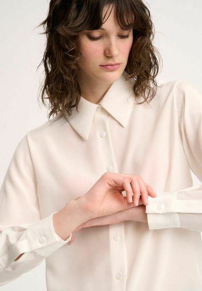 Long-sleeve, button-up shirt in light cream fabric. Features a classic collar, cuffs with buttons, and slightly sheer texture.