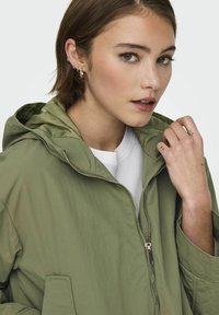Veste à capuche légère de couleur olive, avec une fermeture éclair à l'avant, dotée de deux poches latérales et d'une texture lisse et mate.