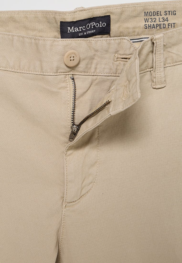 Beżowe bawełniane spodnie z zamkiem błyskawicznym, zapięciem na guziki i kieszeniami bocznymi. Posiadają metkę z nazwą marki i szczegółami rozmiaru.