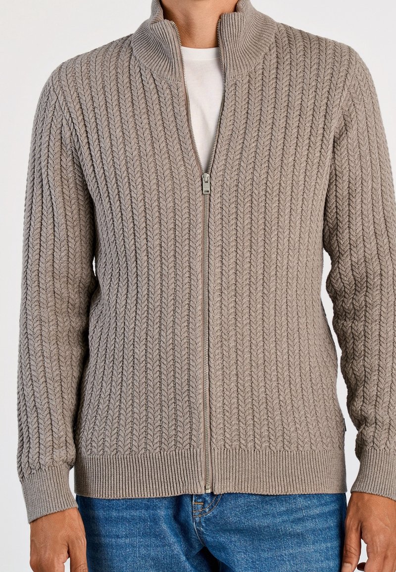 Homme portant un cardigan beige en maille torsadée zippé par-dessus une chemise blanche et un jean bleu, debout devant un fond uni clair.