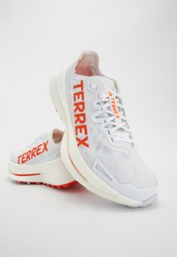 Białe buty sportowe z jaskrawą pomarańczową marką "TERREX", siateczkową cholewką, teksturowaną powierzchnią oraz trwałą białą podeszwą z czerwonymi akcentami.