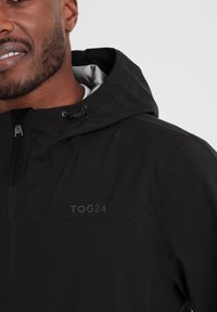 Black waterproof jacket with a hood, featuring a logo print, front zip, and a textured fabric. Includes gray lining for insulation.