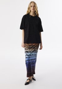 Černé oversized tričko s ozdobami z perel, spárované s dlouhou, vícebarevnou sukní s gradientovým vzorem a potiskem textu.