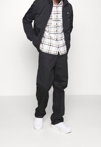 Man som bär svart jacka, vit rutig skjorta, svarta byxor och vita sneakers står mot en enfärgad ljus bakgrund.