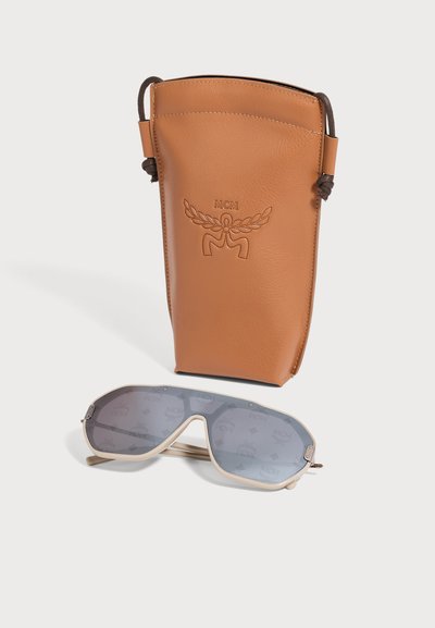 Pochette en cuir marron MCM à côté de lunettes de soleil à monture beige avec des verres gris, les deux posés sur une surface blanche.