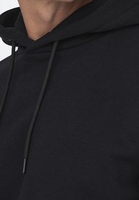 Sudadera negra con capucha de textura suave, que cuenta con un bolsillo frontal tipo canguro y cordones ajustables. La tela parece ligera y casual.
