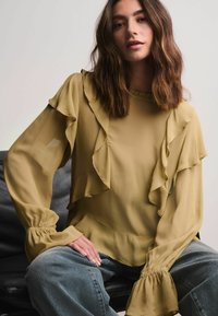 Bluză din chiffon de culoare verde olive, cu detalii cu volane pe decolteu și pe mâneci. Mâneci lungi și fluidi, cu manșete gathered.