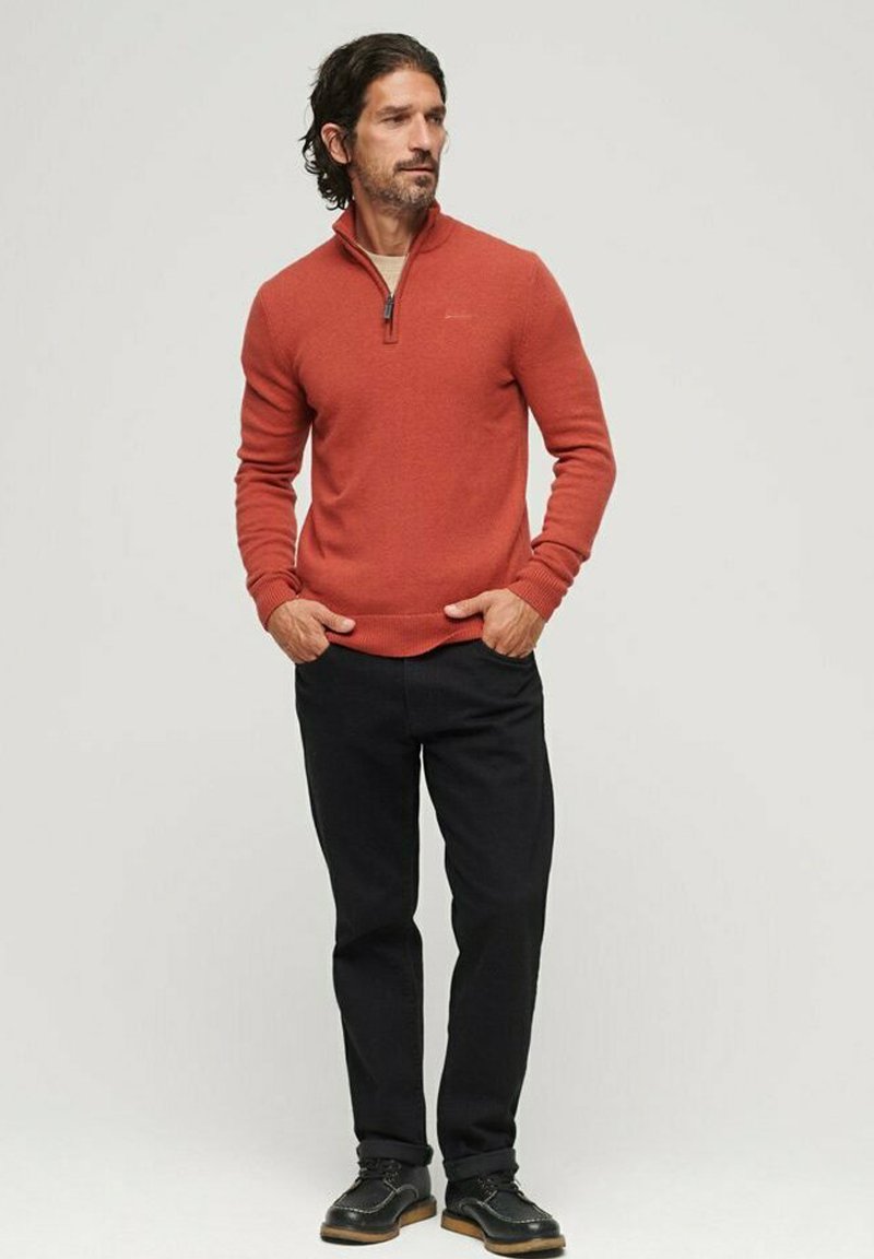 Pull à col zippé orange avec des poignets et un ourlet côtelés, associé à un pantalon noir et des chaussures noires avec une semelle marron clair.