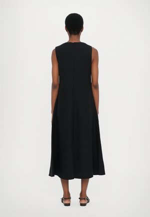 Femme debout de dos, portant une robe midi noire sans manches et des sandales noires ouvertes à l'arrière sur fond blanc.