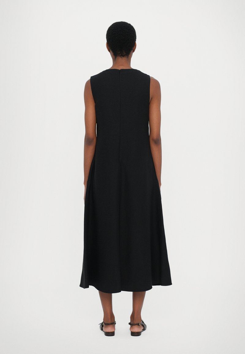 Femme debout de dos, portant une robe midi noire sans manches et des sandales noires ouvertes à l'arrière sur fond blanc.