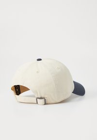 Beige katoenen pet met een marineblauwe rand, verstelbare band en een contrasterende bovenknop. Voorzien van ventilatiegaatjes en een logolabel aan de binnenkant.
