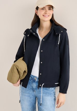 Lächelnde Frau in marineblauer Jacke, weißem Hemd und beiger Mütze hält eine gefaltete olivgrüne Jacke und steht vor einem schlichten hellen Hintergrund.