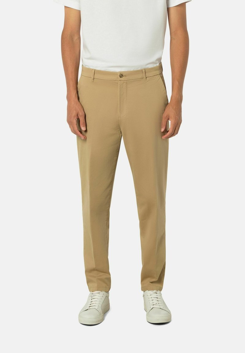 Mann in beigen, schmal geschnittenen Hosen, weißen Turnschuhen und einem weißen Kurzarmhemd, das vor einem einfarbigen Hintergrund steht.