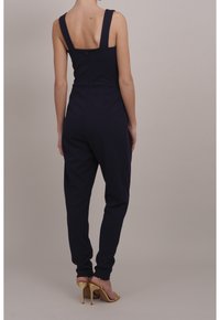 Kvinna i en ärmlös marinblå jumpsuit med breda axelband och dragkedja i ryggen, kombinerad med guldiga högklackade sandaler, stående mot en enfärgad bakgrund.