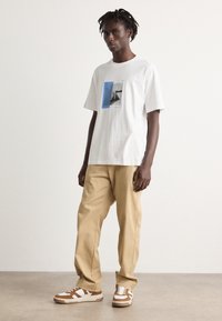 Hvit bomulls t-skjorte med et blått grafisk trykk på fronten, sammen med lysebeige bukser og brune og hvite sneakers, med et sporty design.
