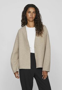 Cardigan beige, surdimensionné, avec un col en V, deux boutons et des manches larges, porté sur un haut blanc, associé à un pantalon noir.