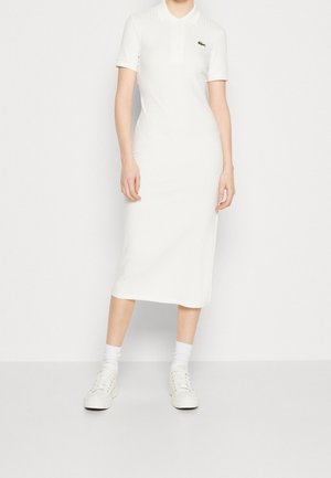 Kvinde iført hvid ribbet midi polo kjole, hvide sokker og hvide sneakers, stående mod en ensfarvet hvid baggrund.