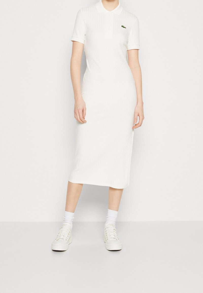 Femme portant une robe polo midi blanche côtelée, des chaussettes blanches et des baskets blanches, debout devant un fond blanc uni.