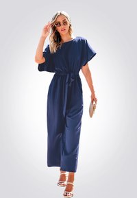 Robe maxi bleu marine à manches courtes avec ceinture à nouer, présentant un design fluide et une texture subtile. Portée avec des sandales de couleur claire et des lunettes de soleil.