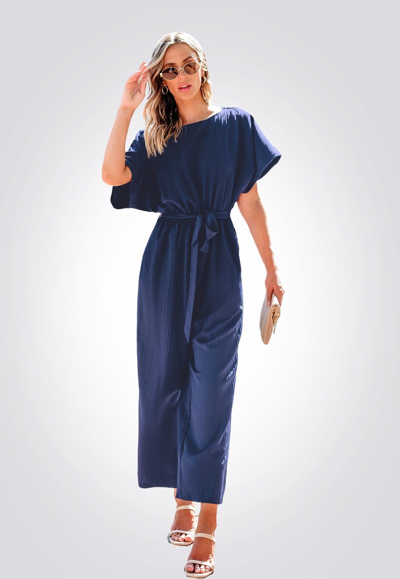 Robe maxi bleu marine à manches courtes avec ceinture à nouer, présentant un design fluide et une texture subtile. Portée avec des sandales de couleur claire et des lunettes de soleil.