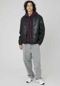 Veste en simili cuir noir avec des poignets élastiques, portée sur un hoodie bordeaux. Le modèle porte un jean baggy gris clair et des chaussures de sport blanches.