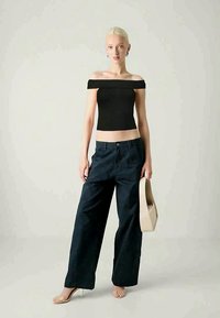 Juodas pečių nuleistas megztinis, tamsiai mėlynos aukšto juosmens plačiakelnės kelnės, neutralus rankinukas ir kūno spalvos kulniniai sandalai prieš šviesų foną.