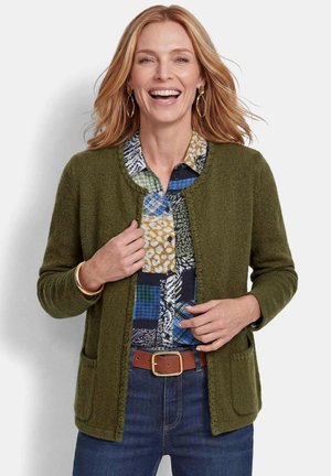 Lächelnde Frau mit blondem Haar trägt grünen Strickcardigan über gemustertem Hemd, blaue Jeans, braunen Gürtel, goldene Ohrringe und Armband.