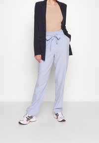 Person som bär ljusblå paperbag midja byxor, beige topp, svart randig blazer och vita sneakers står mot en vit bakgrund.