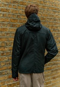 Jachetă cu capușon neagră, realizată dintr-un material neted și ușor. Are un design simplu, cusături vizibile și o croială dreaptă, purtată peste pantaloni bej.