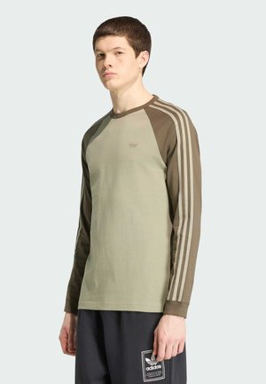 Ung mand iført en grøn Adidas langærmet trøje med tre striber på ærmerne og sorte Adidas bukser med logo-patch.