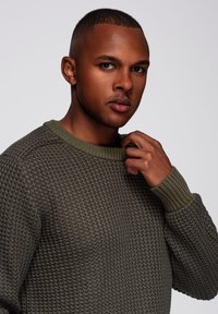 Suéter texturizado de color verde oliva con un cuello redondo de canalé y puños de canalé. La tela presenta un patrón de panal y un diseño ligeramente ajustado.