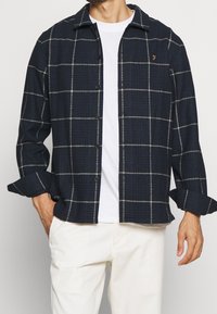 Camicia a quadri blu navy con linee bianche e grigie, maniche lunghe e taschino sul petto; indossata sopra una maglietta bianca.