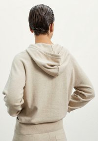 Felpa beige con una texture morbida, caratterizzata da un cappuccio con apertura sul retro e polsini e orlo a coste. Il design è semplice e aderente.