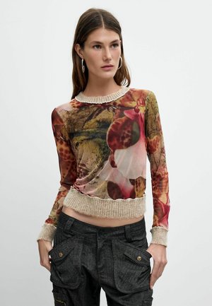 Kvinde med langt brunt hår iført øreringe, en gennemsigtig, blomsterprintet bluse med lange ærmer med beige strikkede manchetter og mørke cargobukser.