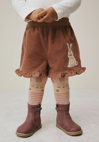 Barn iført brune rynkede shorts med kaninapplikation, stribede kaninansigtstrømpebukser og matchende brune ankelstøvler, stående på lys gulv.