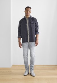Mörkgrå kordjacka med ficka framtill, bärs över en ljus rutig skjorta. Ljusblå jeans och grå sneakers fulländar outfiten.
