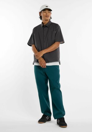 Mann trägt weiße Kappe, dunkelgraues kurzärmeliges Hemd mit Knöpfen, türkisfarbene Hose und schwarze Turnschuhe, steht vor weißem Hintergrund.