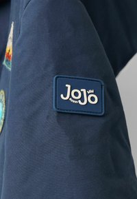 Jūras spēku audums ar teksturētu virsmu, ar zilu taisnstūra etiķeti, uz kuras baltajās paceltajās burtiņās ir uzraksts "JoJo bébe", kā arī dažādas izšūtas uzšuvumi.