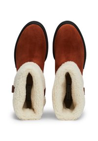 Bruine suède enkellaarsjes met een fluffy crèmekleurige schapenvacht kraag en een zwarte zool, met een detail van een zijband. De binnenkant is gevoerd met zacht materiaal.